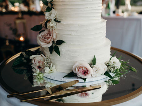 A white wedding cake with flowers and golden utensils, sitting on a reflective platter.