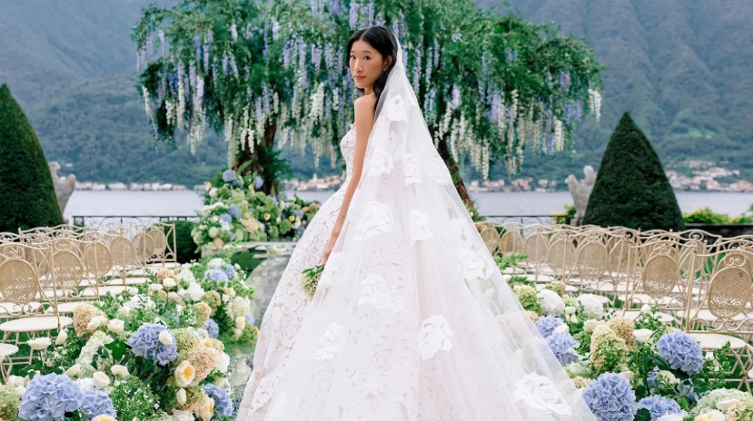 Bride in a floral lace gown stands in a scenic outdoor setting with mountains, flowers, and empty chairs, exuding elegance and serenity.
