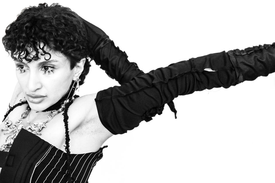 High-contrast portrait of a person with short curly hair, long black gloves and pinstriped garment, arms raised – Photography from the series 'Transgression durch Hoffnung' by Alfred Stoll, 2025–2026. Metamodern Art; Exhibition kultur.lokal.fürth; Metamodernism; Metamodern Grotesk.