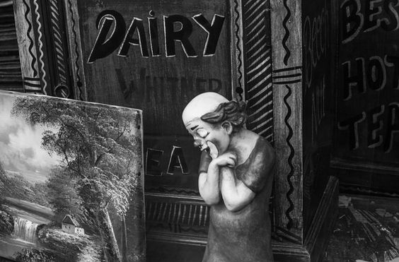 Sad clown figurine in front of wooden cabinet with hand-painted signs reading 'DAIRY' and 'TEA', small landscape painting beside it – Photography from the series 'Transgression durch Hoffnung' by Alfred Stoll, 2025–2026. Metamodern Art; Exhibition kultur.lokal.fürth; Metamodernism; Metamodern Grotesk.