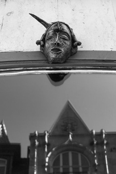 Weathered mask with closed eyes above reflective surface, horn-like protrusion, building reflection below – Photography from the series 'Transgression durch Hoffnung' by Alfred Stoll, 2025–2026. Metamodern Art; Exhibition kultur.lokal.fürth; Metamodernism; Metamodern Grotesk.