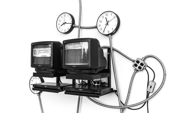 Media installation with two CRT televisions, DVD players and three analog clocks connected by flexible metal conduits on a white wall – Photography from the series 'Transgression durch Hoffnung' by Alfred Stoll, 2025–2026. Metamodern Art; Exhibition kultur.lokal.fürth; Metamodernism; Metamodern Grotesk.