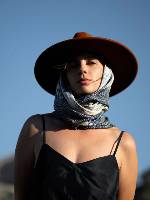 Outdoor portrait of a woman in a wide-brim hat and patterned scarf, photographed from a low angle against a clear blue sky