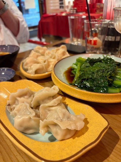 Dumplings and greens in soy sauce on plates, with drinks and glasses on a wooden table in a cozy dining setting. Vibrant reds in background.