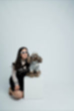 Woman kneeling next to a fluffy dog, both wearing heart-shaped sunglasses. Dog in patterned outfit, set against a plain light background.