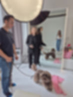 Man holding a leashed dog in a studio, woman in black suit stands nearby, mirror reflects another woman. Pink polka-dot cloth on floor.