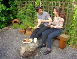 A couple are drinking tea and eating cake on a bench in a secluded garden