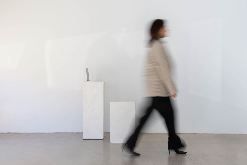 A motion-blurred image of a woman walking past white pedestals and a laptop in a minimalist studio.