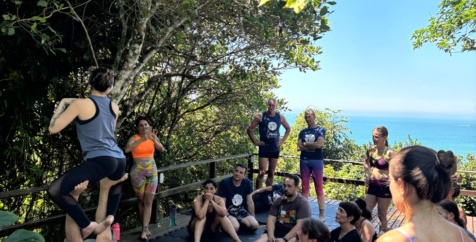 Dupla ensinando acroyoga para turma de frente para o mar em meio a natureza e céu azul