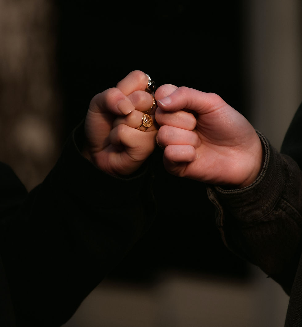 Two hands in a fist bump, both wearing black sleeves. One hand has two gold rings. Dark background, conveying a friendly gesture.