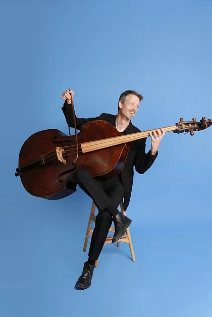 Malachy Robinson seated on a stool with his double bass, against a blue background.