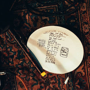 A paper plate with the Westside Cowboy setlist written in black Sharpie, resting on a patterned carpet. A guitar pedal peeks out from the bottom left corner.
