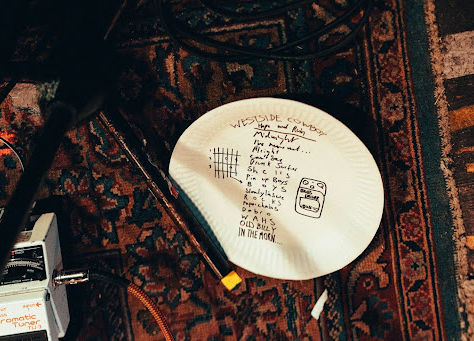 A paper plate with the Westside Cowboy setlist written in black Sharpie, resting on a patterned carpet. A guitar pedal peeks out from the bottom left corner.