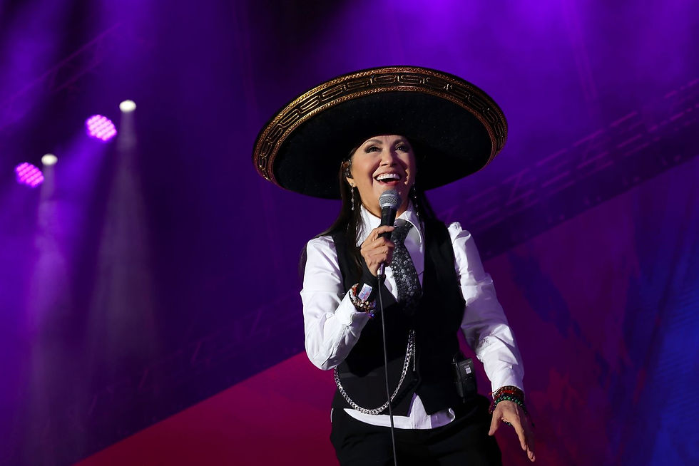 Cantora Ana Gabriel sorridente de sombrero canta ao microfone em palco iluminado de roxo.