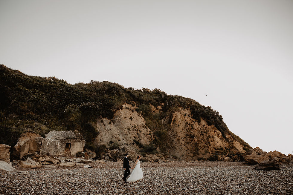 Koppelshoot van bruidspaar dat gaat trouwen in Frankrijk, terwijl ze tijdens hun elopement samen over een kiezelstrand wandelen met rotsen op de achtergrond - huwelijksfotograaf Gent