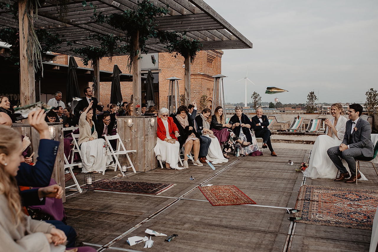 Gasten gooien papieren vliegtuigjes naar het bruidspaar tijdens een persoonlijke ceremonie op het dakterras van trouwlocatie rooftopbar Gaston in Gent