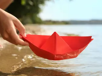 Close-up of hand placing red paper boat at water's edge, representing the first step in your wealth planning journey