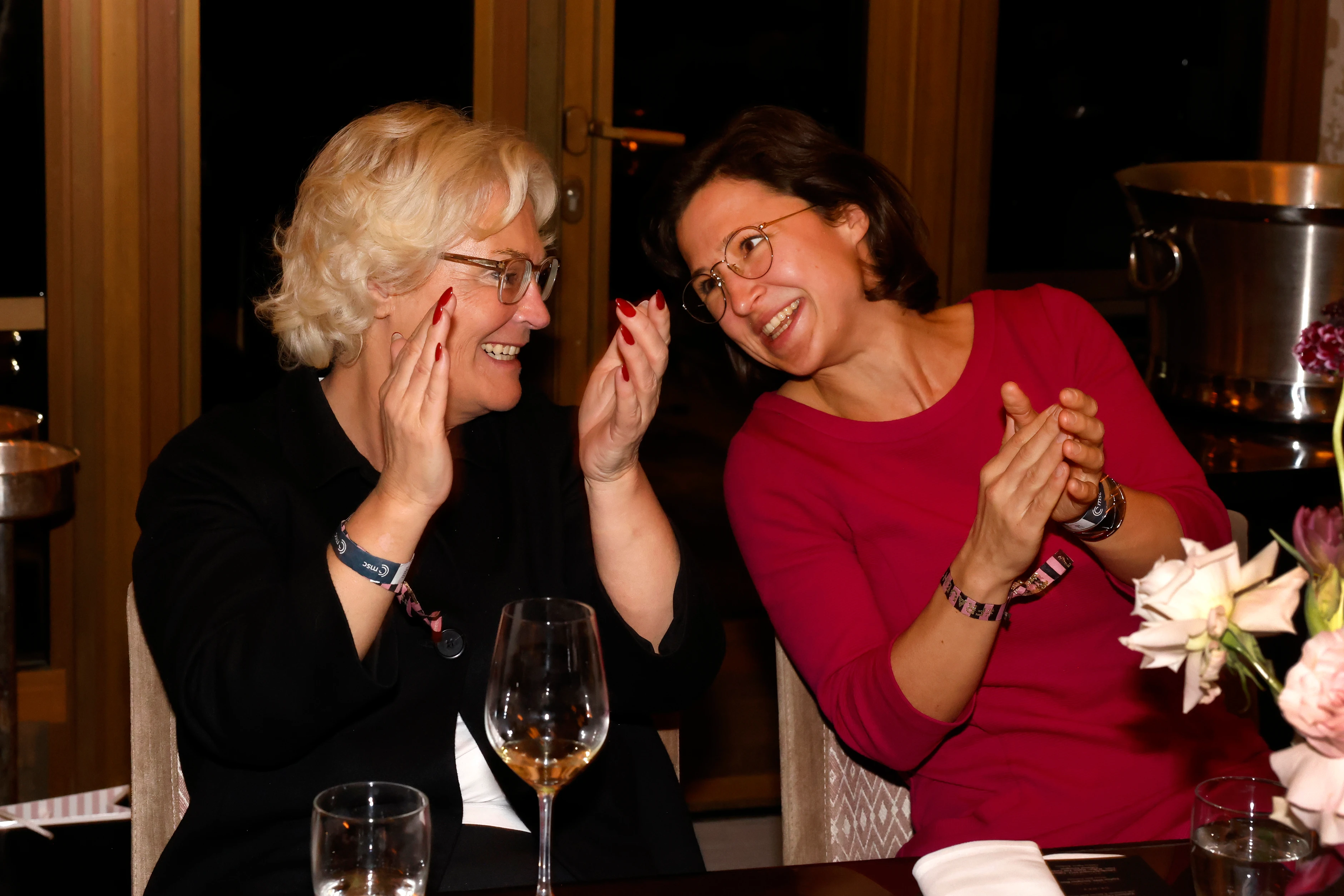 Zwei Frauen sitzen nebeneinander an einem Tisch, lachen miteinander und applaudieren, während sie sich zugewandt unterhalten.