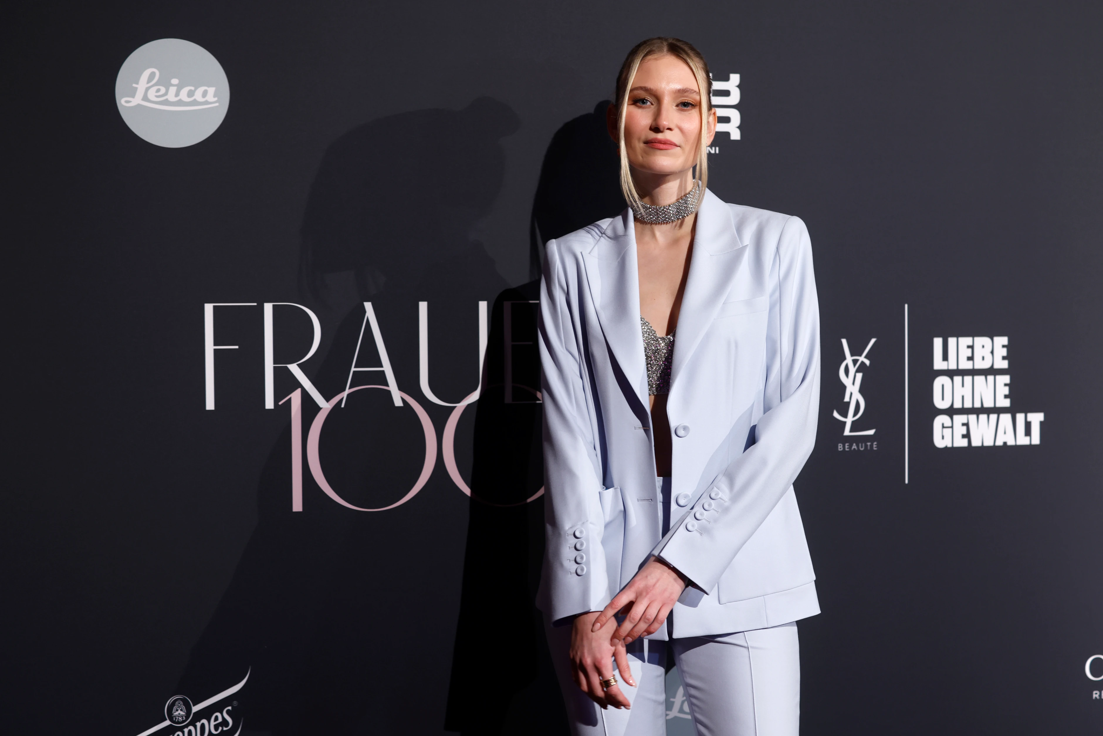 Frau im hellen Hosenanzug posiert vor „Frauen100“-Eventwand mit Logos und Slogan „Liebe ohne Gewalt“.