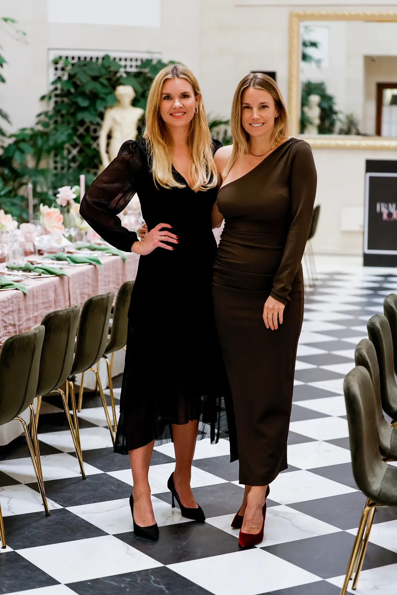 Zwei Frauen stehen in einem elegant dekorierten Raum neben einer langen Tafel und posieren für ein Foto.