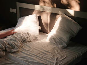 Unaufgeräumtes Bett mit zerknitterter Bettdecke und Kissen, durch das warmes Sonnenlicht fällt; eine Hand ist am Rand der Decke zu sehen.