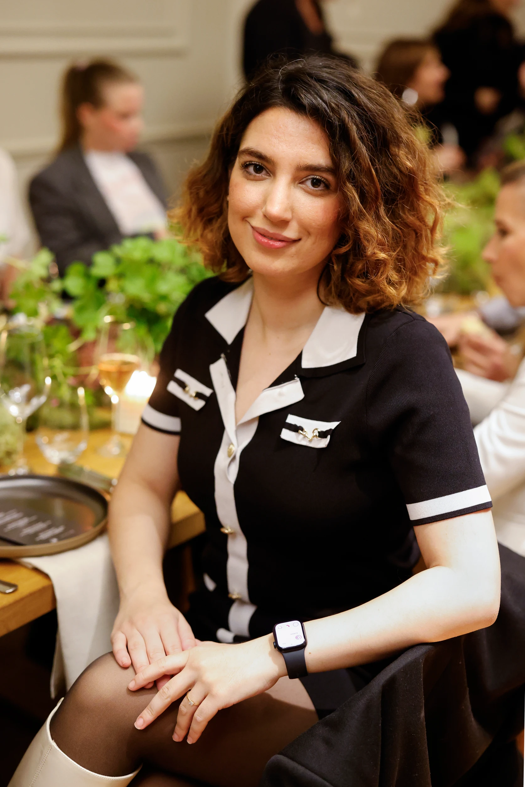 Eine Frau sitzt am Tisch und blickt direkt in die Kamera, während im Hintergrund andere Gäste zu sehen sind.