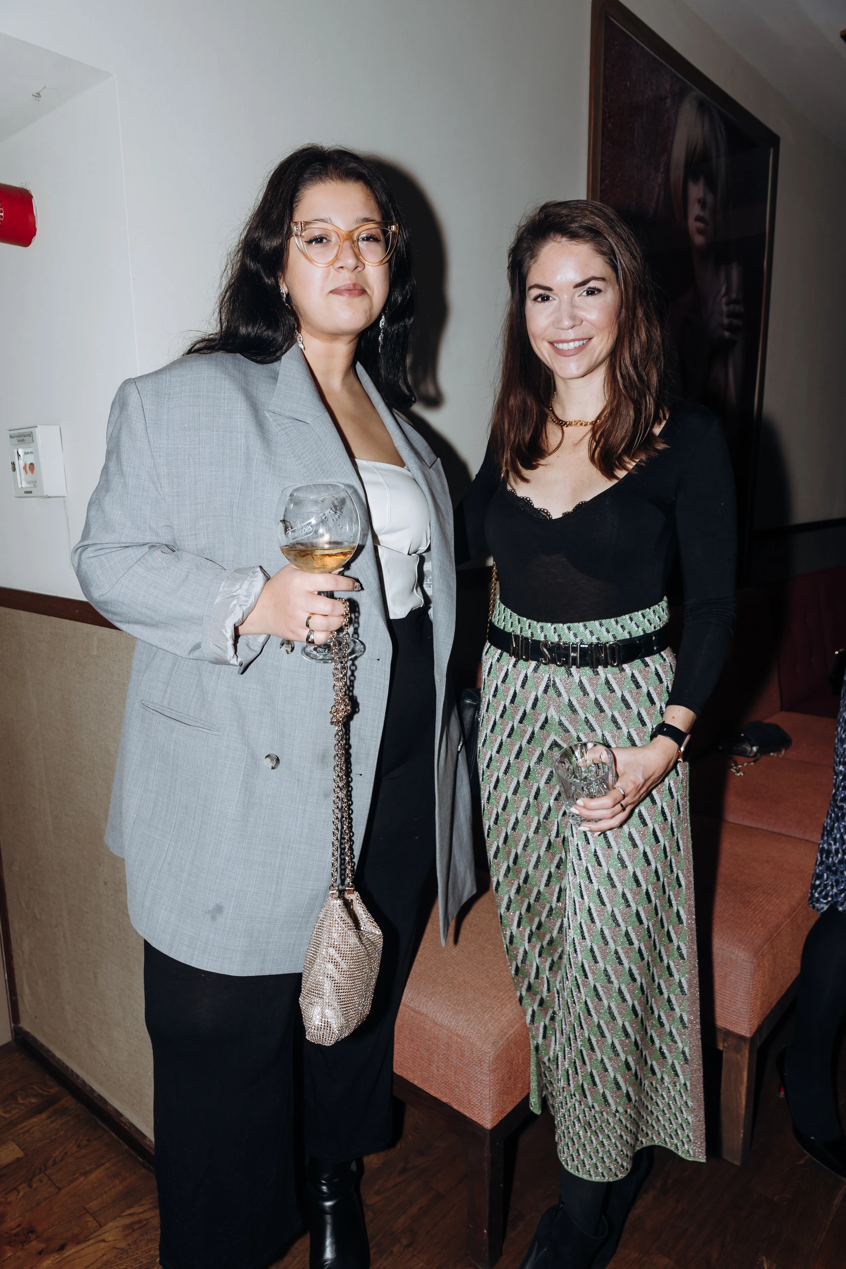 Zwei Frauen stehen nebeneinander bei einem Event, eine im grauen Blazer mit Glas, die andere im schwarzen Oberteil und gemustertem Rock.