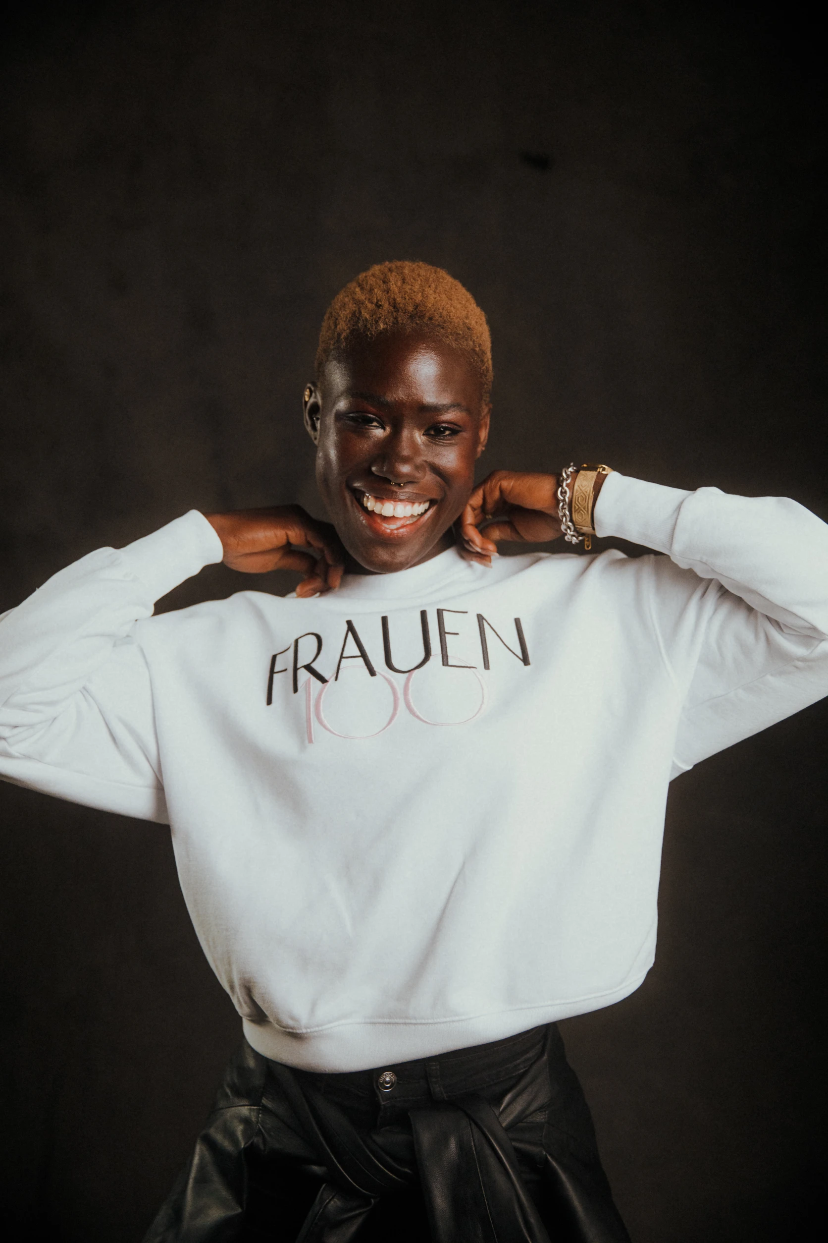 Frau mit kurzen blonden Haaren trägt weißes „Frauen100“-Sweatshirt und lächelt strahlend in die Kamera.