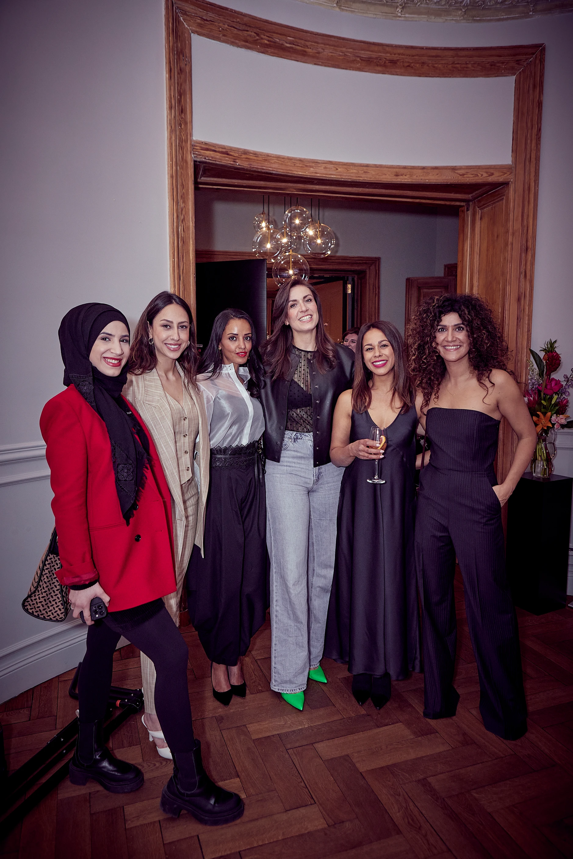 Sechs Frauen stehen eng nebeneinander, lächeln in die Kamera und posieren gemeinsam in einem Raum mit Holzrahmen und warmem Licht.