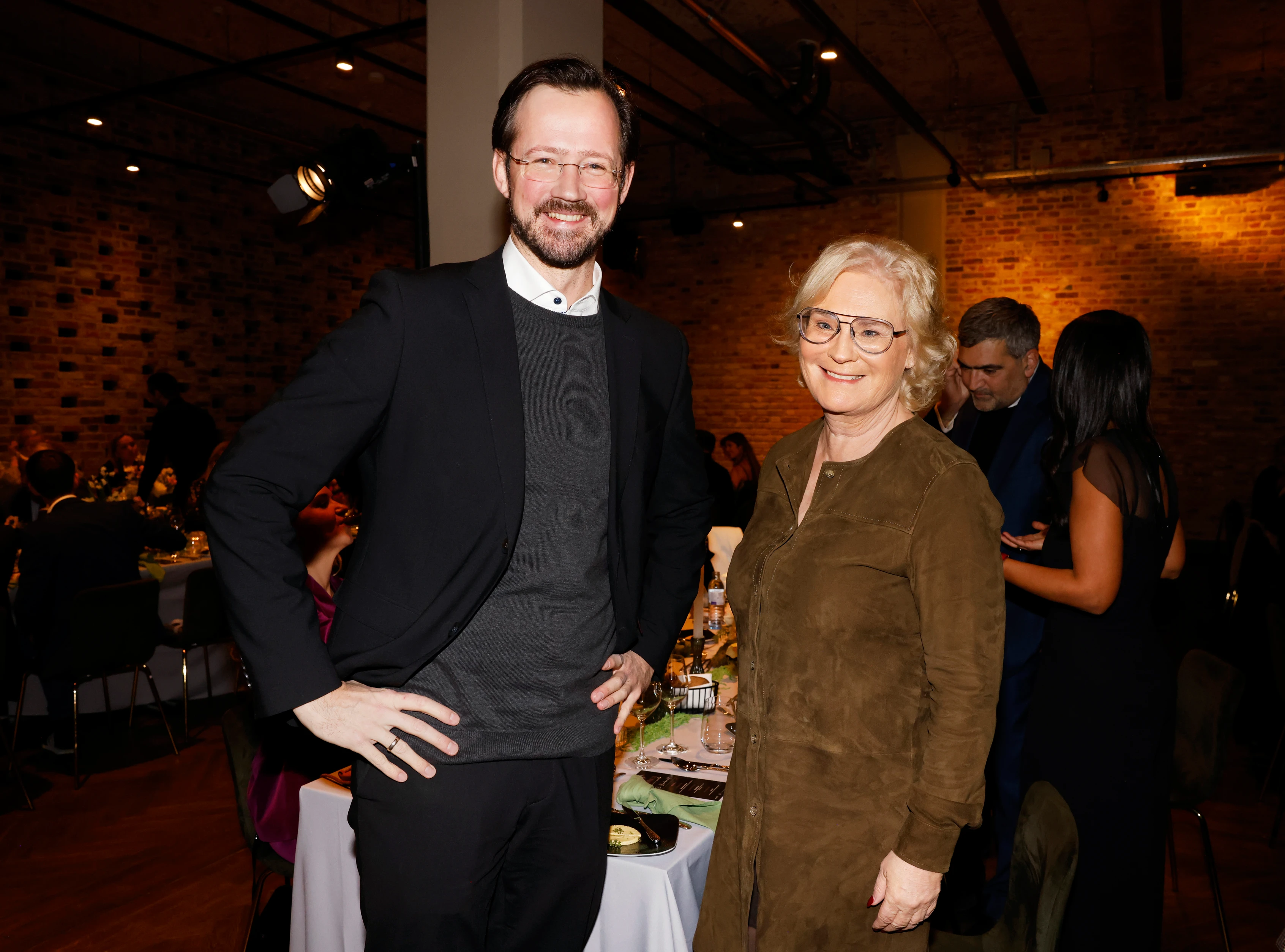 Ein Mann und eine Frau stehen lächelnd in einem Veranstaltungsraum und posieren gemeinsam vor gedeckten Tischen.