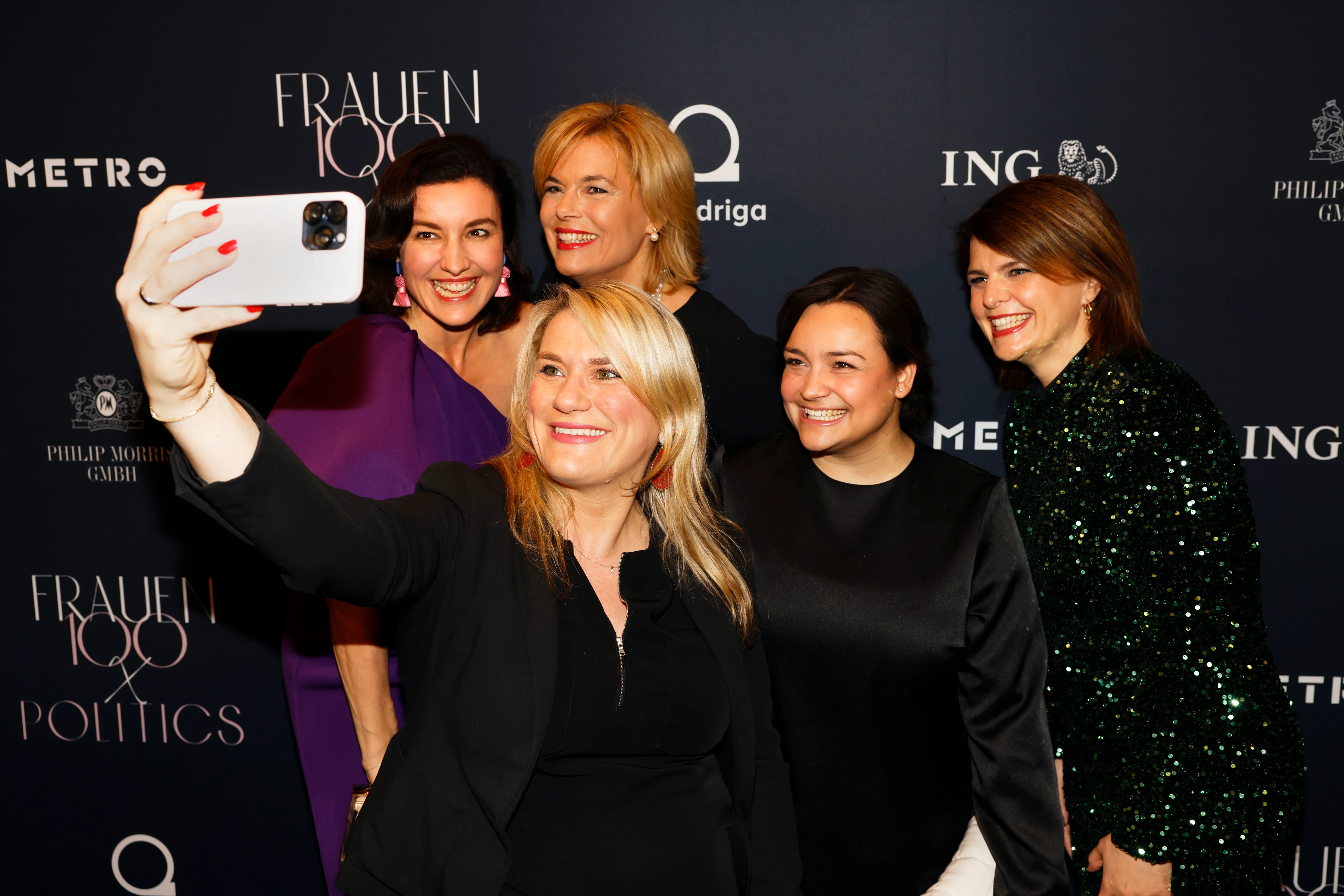 Mehrere Frauen stehen vor einer Event-Wand, lächeln und machen gemeinsam ein Selfie mit einem Smartphone.