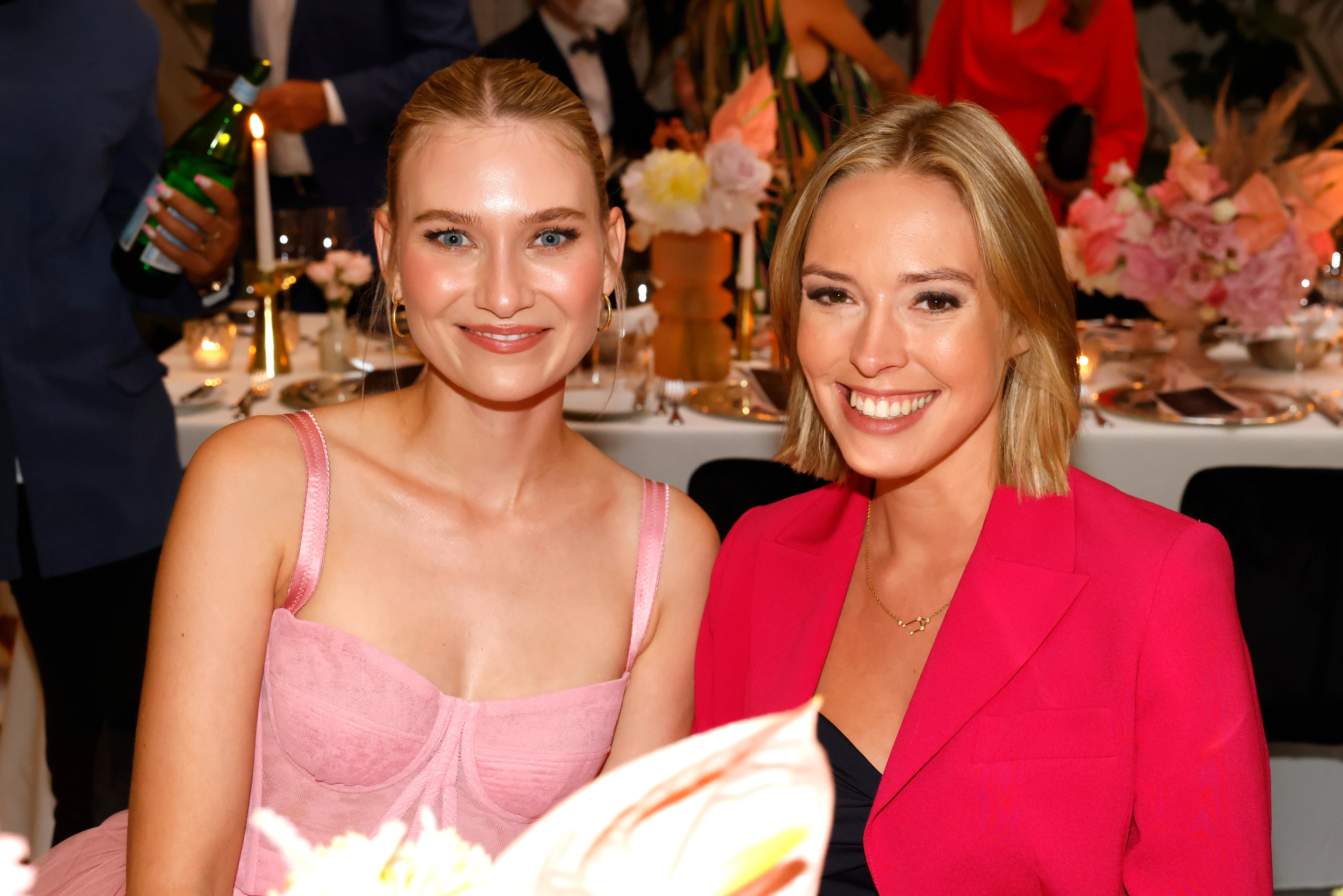 Zwei Frauen sitzen nebeneinander an einem festlich gedeckten Tisch und lächeln in die Kamera, im Hintergrund Kerzen und Blumenarrangements.