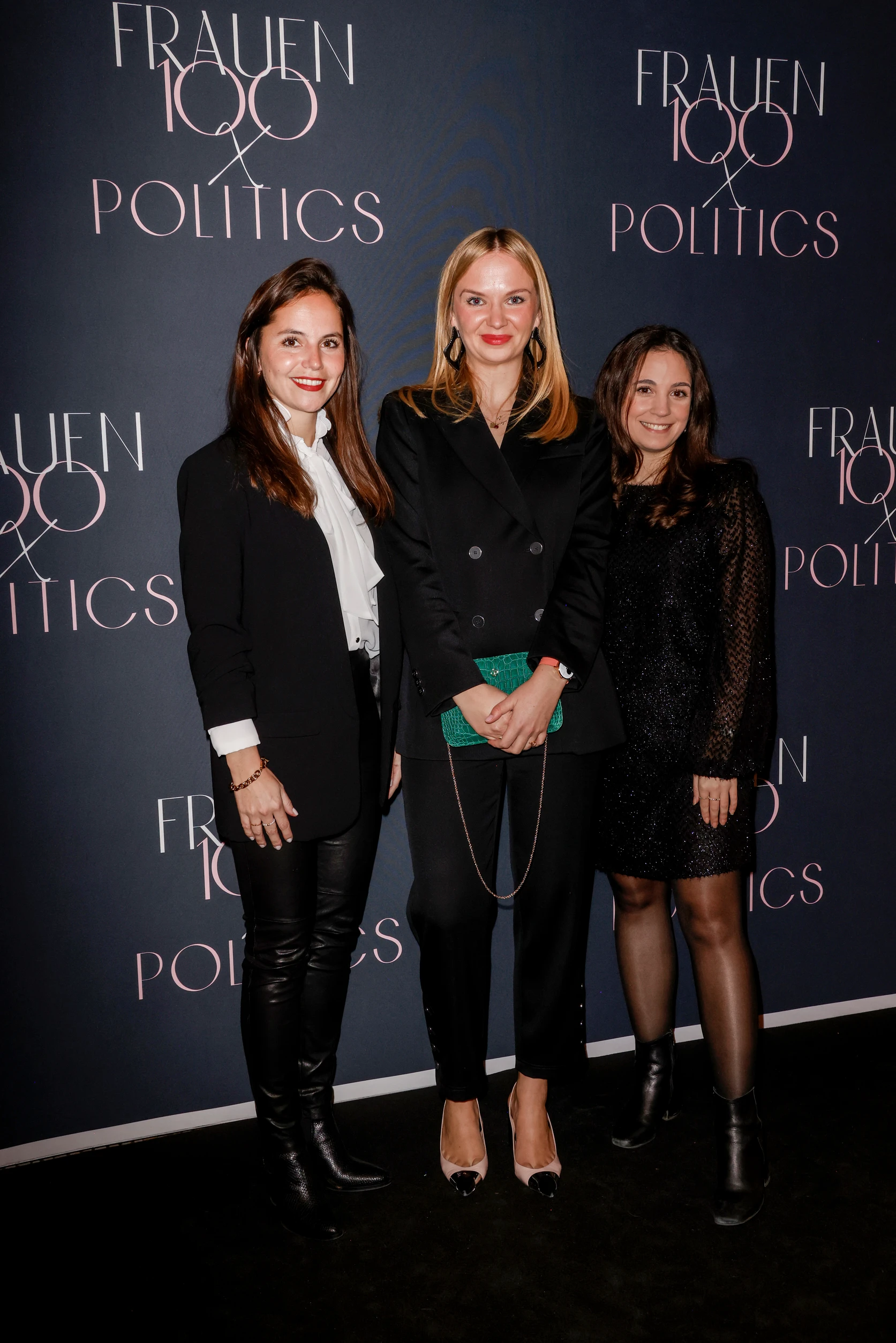 Drei Frauen posieren vor einer Fotowand mit der Aufschrift „FRAUEN 100 & POLITICS“ und lächeln in die Kamera.