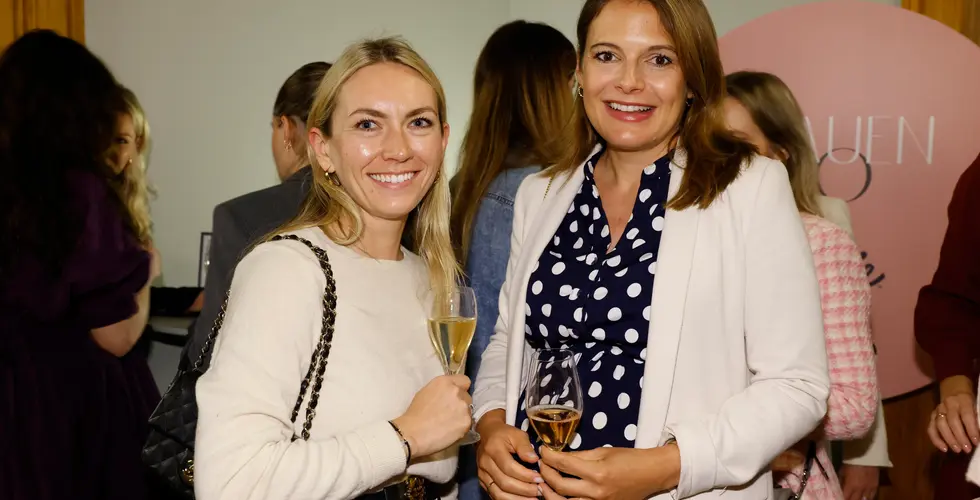 Zwei Frauen stehen eng beieinander, lächeln in die Kamera und halten jeweils ein Glas Sekt in der Hand.