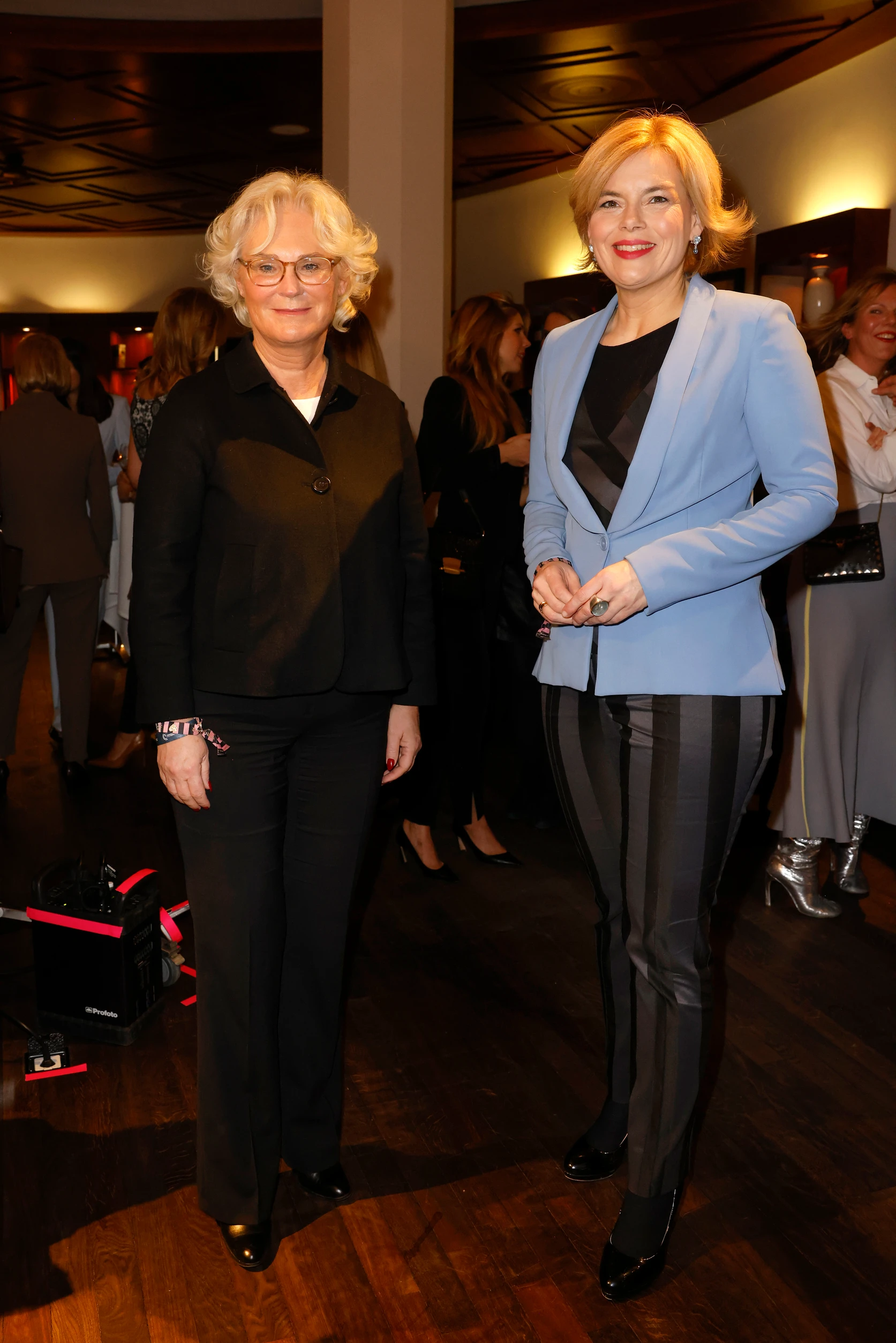 Zwei Frauen posieren stehend für ein Foto, beide elegant gekleidet in einem Raum mit weiteren Gästen im Hintergrund.