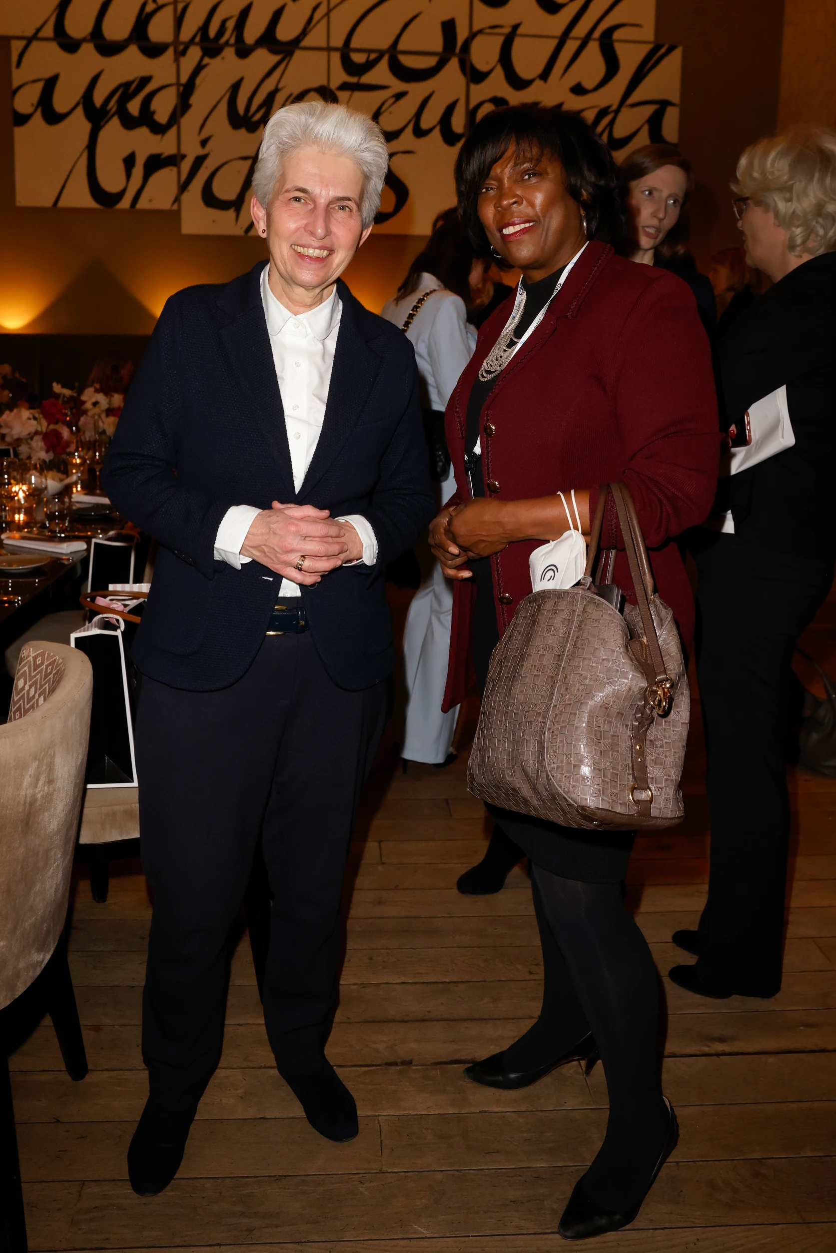 Zwei Frauen stehen nebeneinander, lächeln in die Kamera und halten jeweils eine Handtasche, im Hintergrund ein Event mit Gästen.