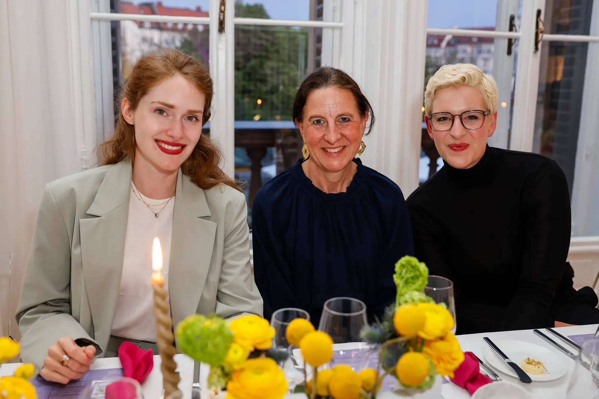 Drei Frauen sitzen gemeinsam am Tisch, lächeln in die Kamera und sind von Kerzenlicht und Blumen umgeben – ein ruhiger, eleganter Moment mitten im Event.
