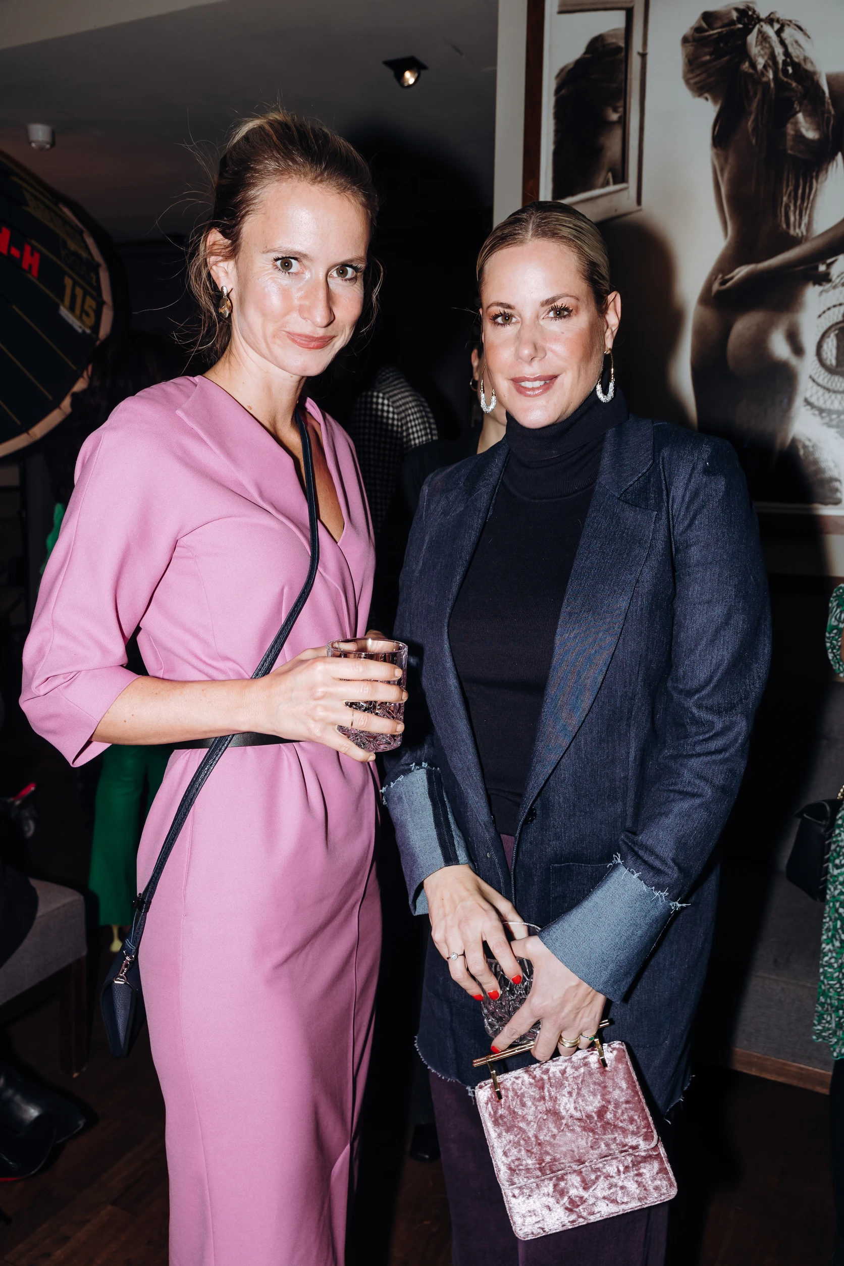 Zwei Frauen posieren bei einem Event, eine im rosa Kleid mit Getränk, die andere im dunklen Blazer mit Handtasche.