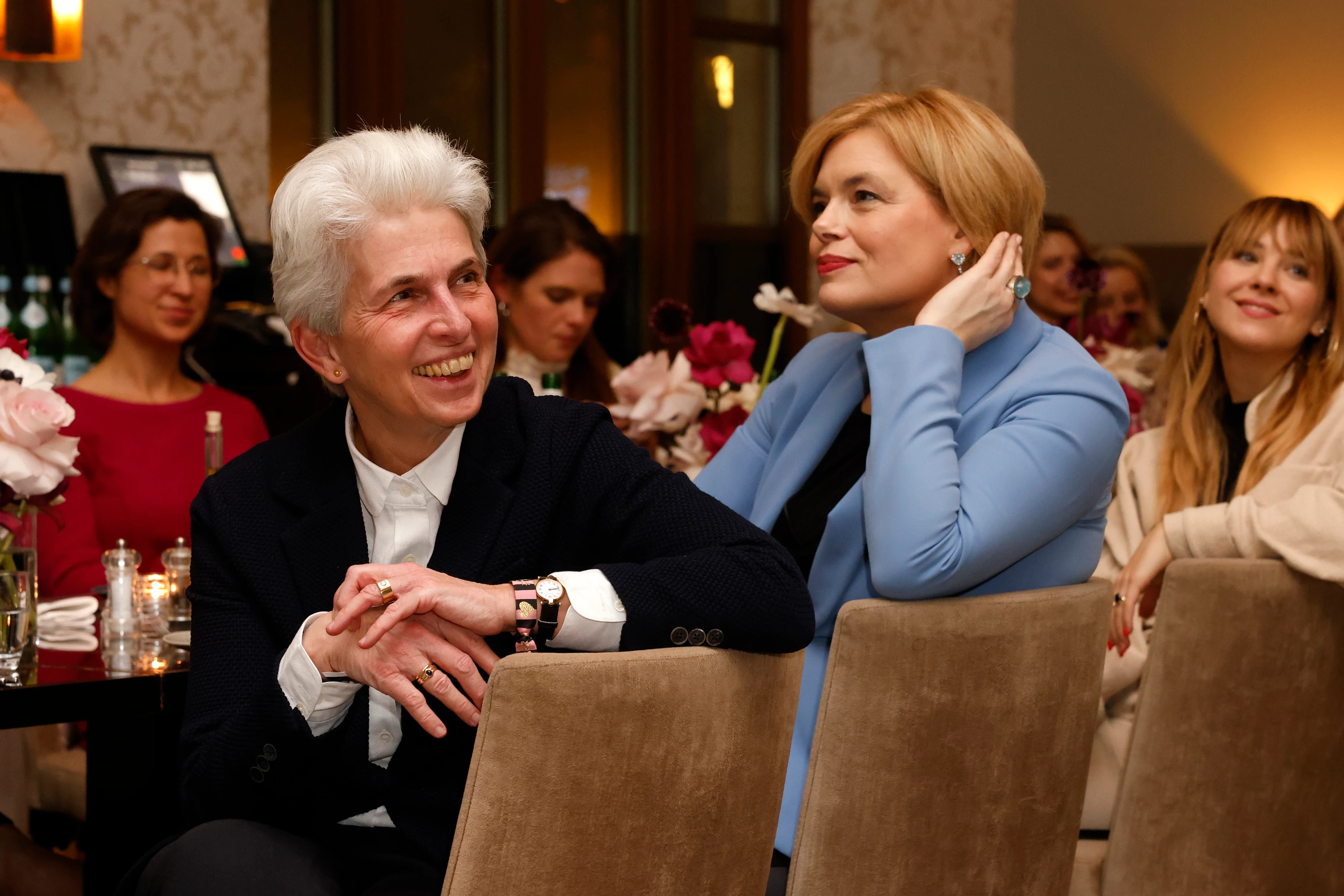 Zwei Frauen sitzen nebeneinander an einem Tisch und hören aufmerksam zu, umgeben von weiteren Gästen und Blumenarrangements.