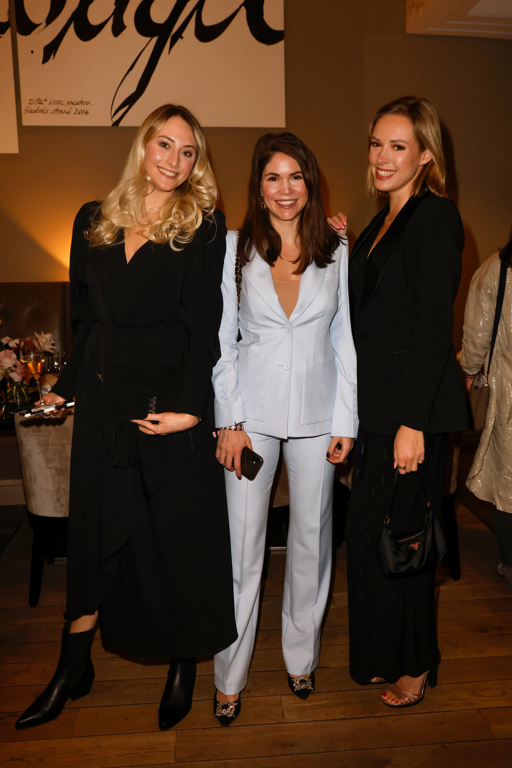 Drei Frauen stehen nebeneinander und lächeln in die Kamera, elegant gekleidet bei einem Event in Innenräumen.