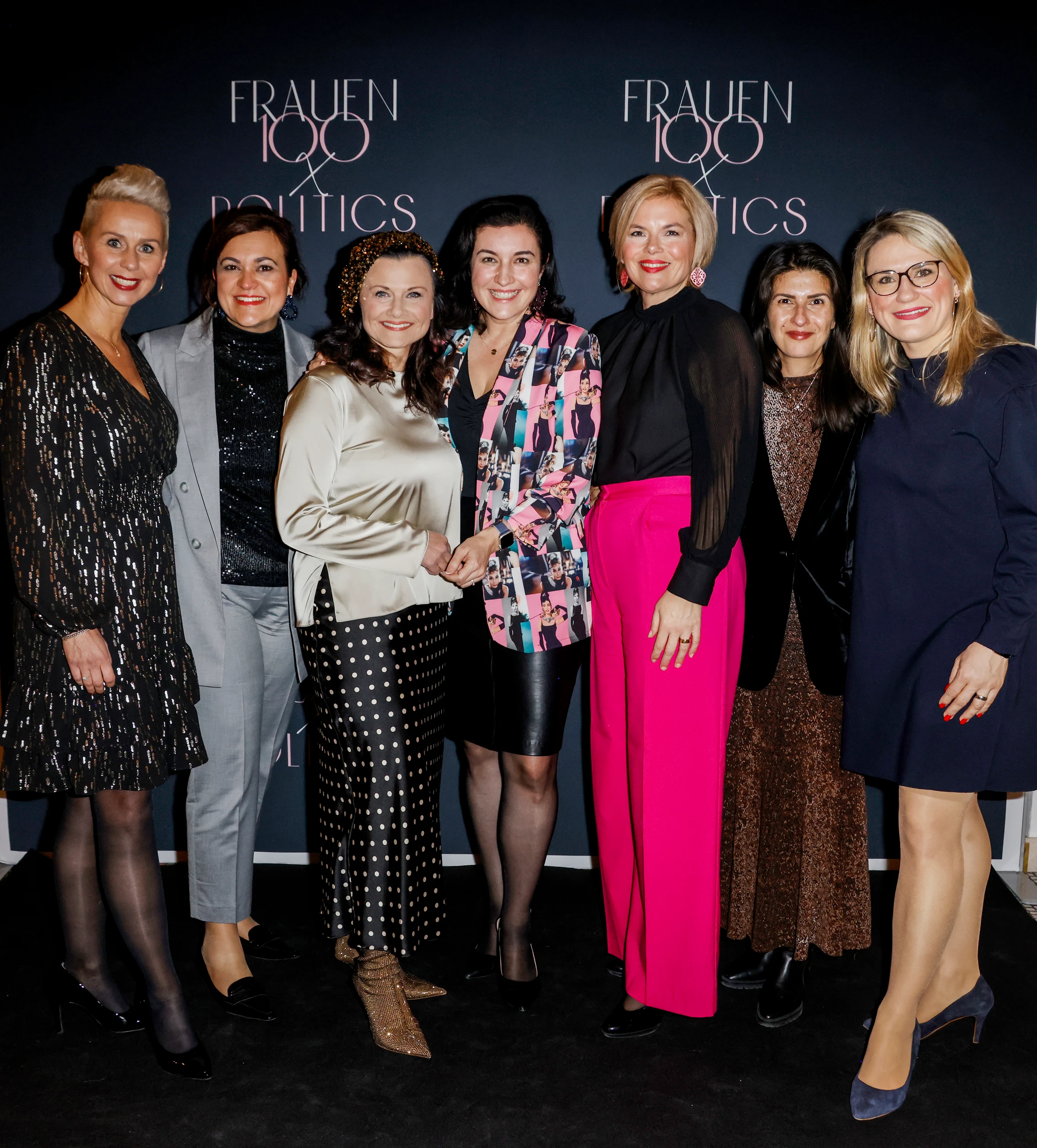 Gruppe von Frauen steht vor einer Fotowand mit der Aufschrift „FRAUEN 100 & POLITICS“ und posiert gemeinsam für ein Foto.