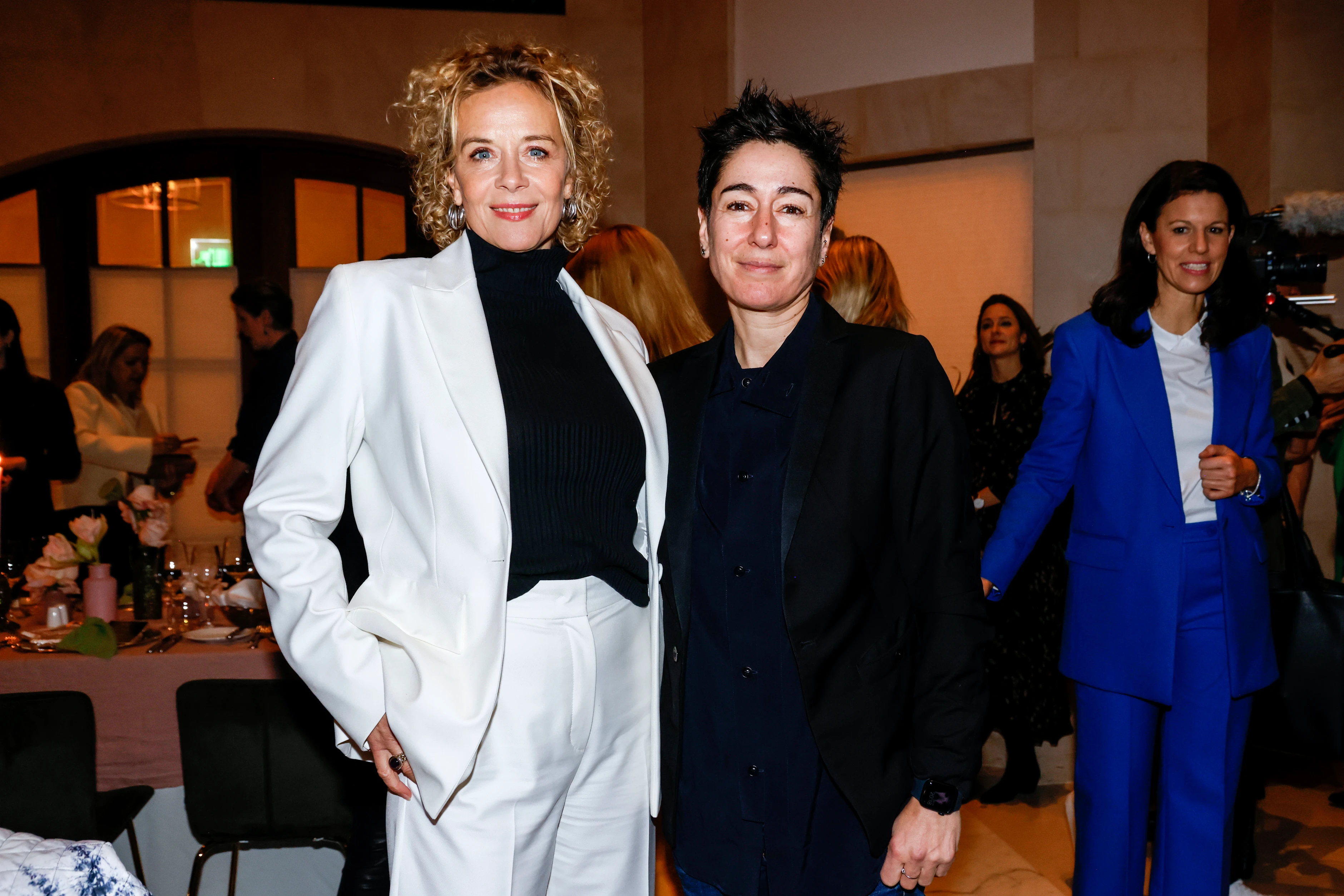 Zwei Frauen stehen bei einem Event nebeneinander und posieren für ein Foto, im Hintergrund weitere Gäste.