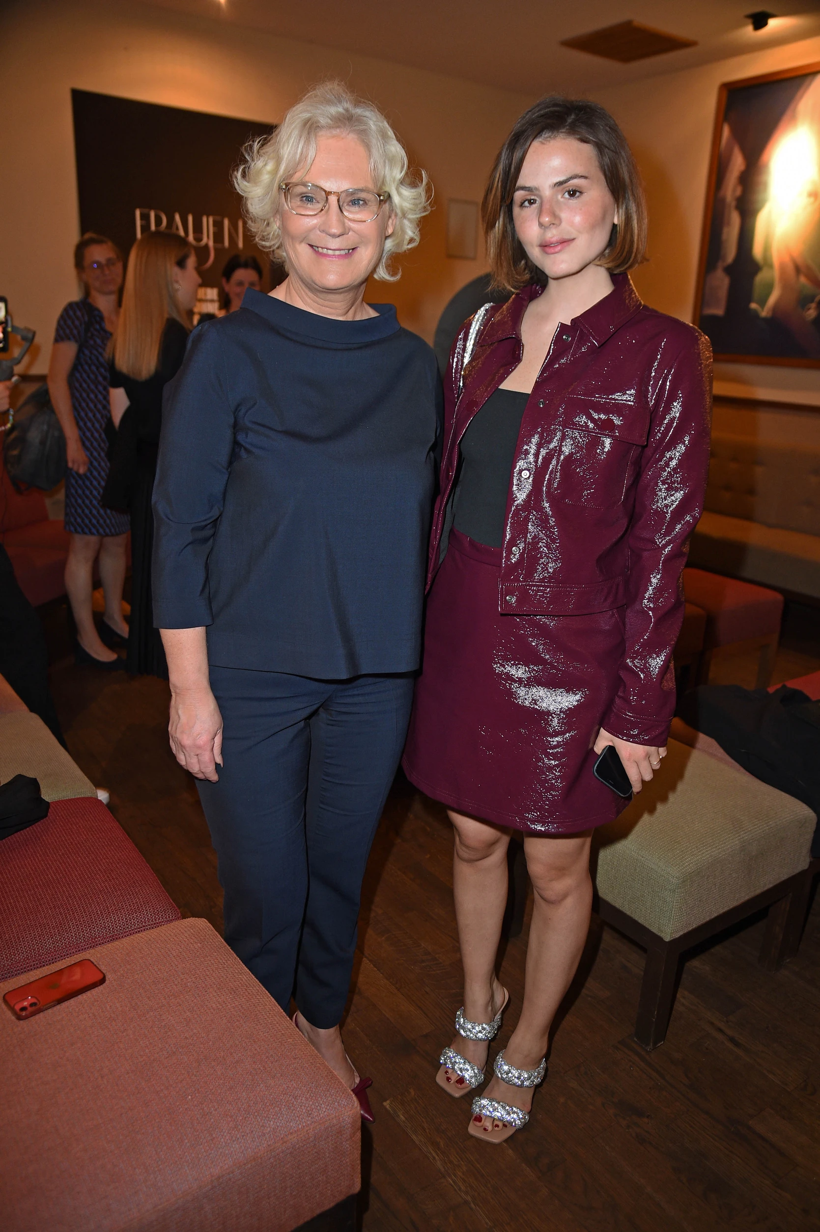 Zwei Frauen stehen nebeneinander bei einem Event und lächeln in die Kamera; eine trägt ein dunkelblaues Outfit, die andere ein glänzendes bordeauxfarbenes Set, im Hintergrund weitere Gäste und warmes Licht.