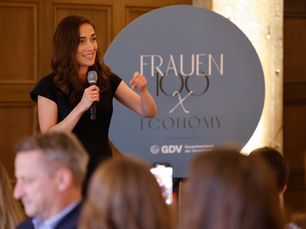 Frau spricht mit Mikrofon vor Publikum auf einer Veranstaltung; im Hintergrund ein rundes Schild mit der Aufschrift „Frauen100 x Iconomy“.