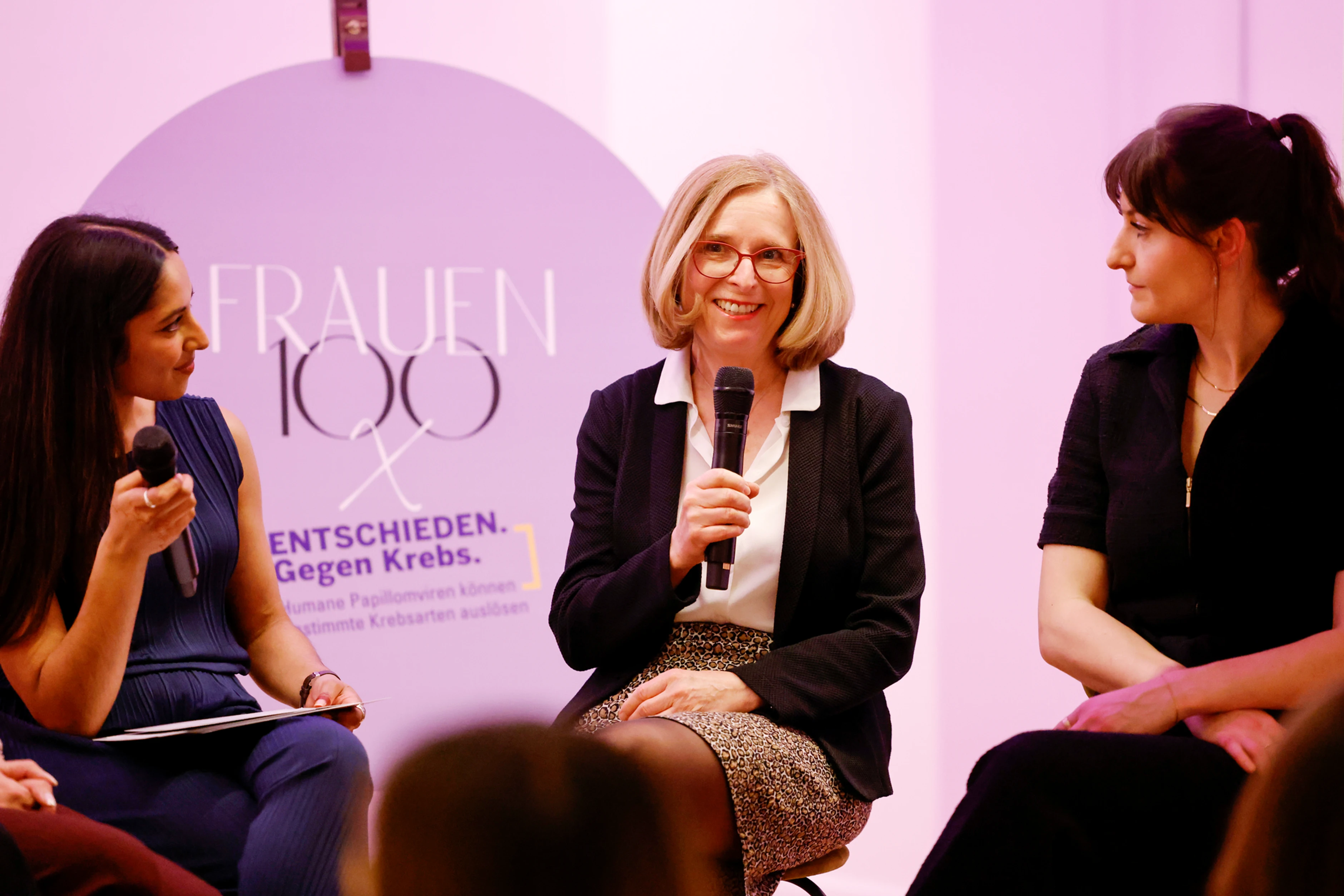 Eine Rednerin spricht ins Mikrofon, während zwei weitere Frauen ihr aufmerksam zuhören – im Hintergrund ist das „Frauen 100“-Branding sichtbar.