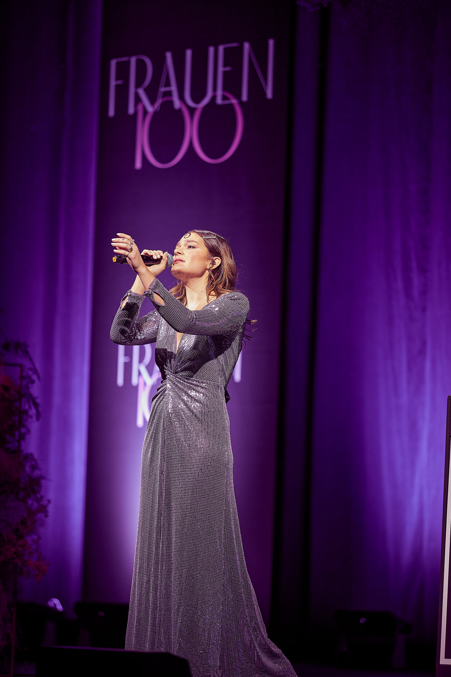 Eine Sängerin im Abendkleid hält ein Mikrofon und performt auf einer Bühne vor einem „FRAUEN100“-Hintergrund.