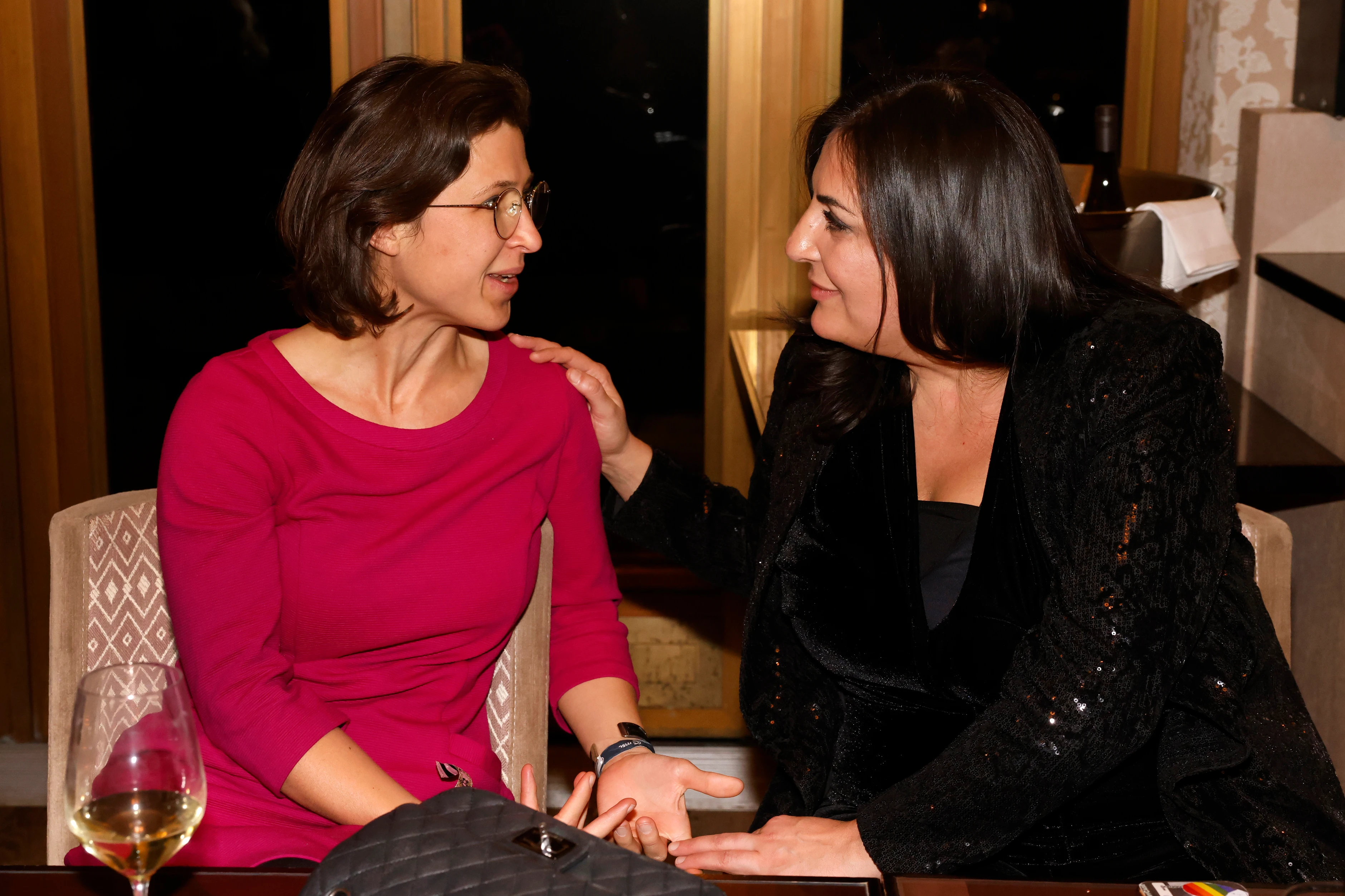 Zwei Frauen sitzen einander zugewandt an einem Tisch, unterhalten sich und lächeln; eine legt der anderen freundschaftlich die Hand auf die Schulter.