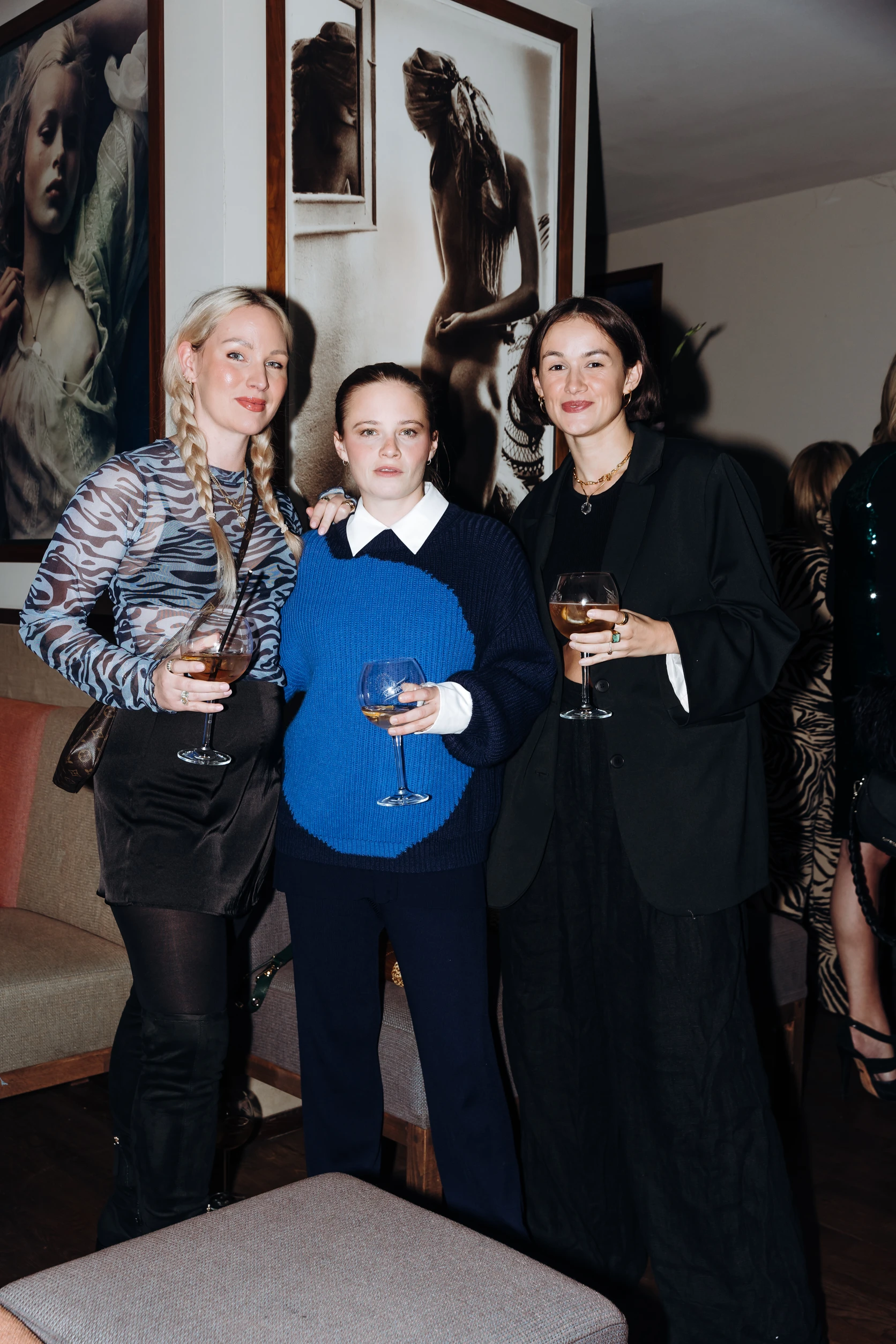 Drei Frauen stehen zusammen mit Getränken in der Hand und schauen in die Kamera, sie tragen elegante Abendlooks.