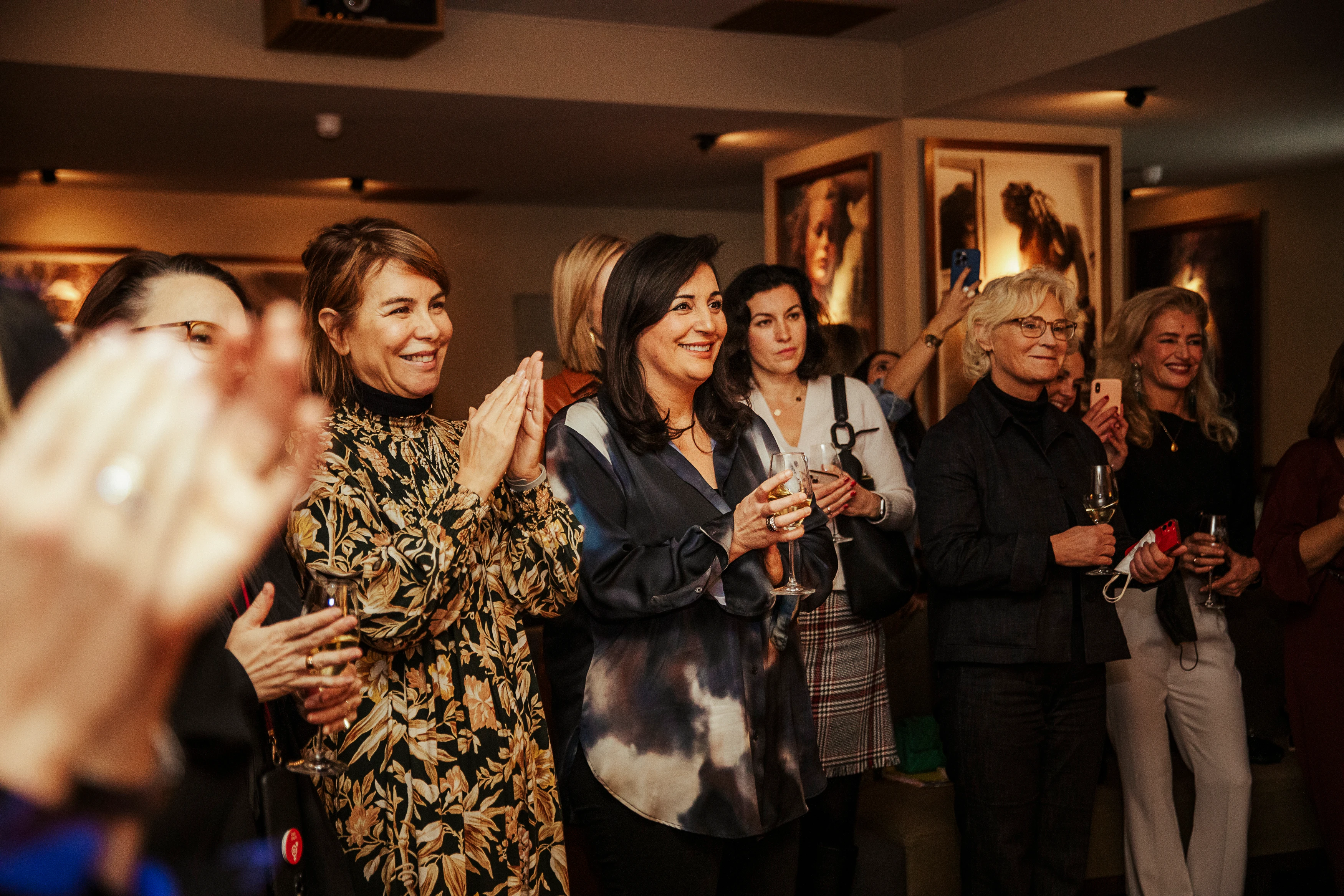 Mehrere Frauen stehen bei einem Event zusammen, halten Getränke in der Hand und applaudieren, während sie aufmerksam in eine Richtung schauen.