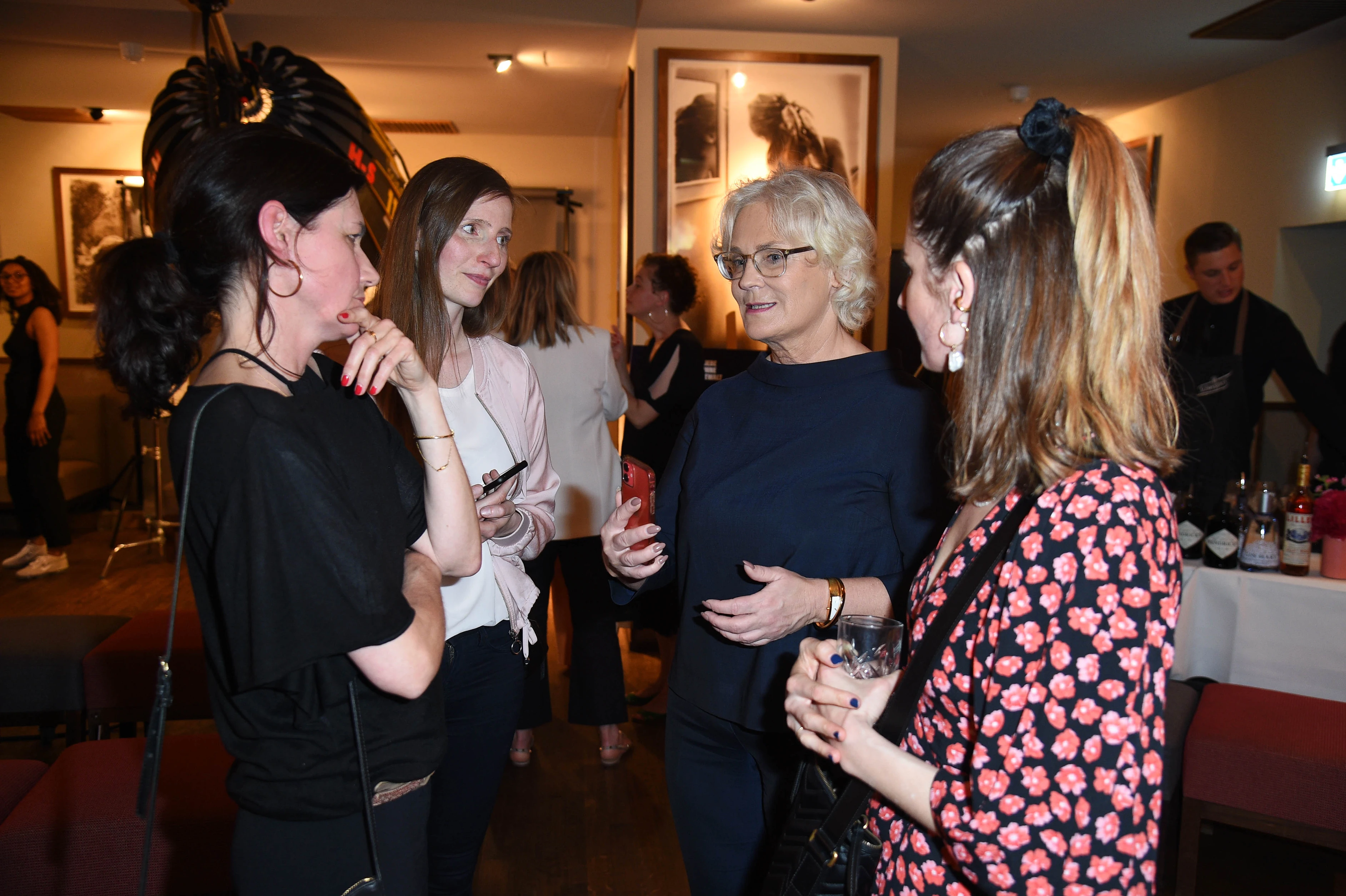 Vier Frauen stehen in einer kleinen Gruppe zusammen und unterhalten sich bei einem Event; sie halten Getränke in der Hand und wirken in ein Gespräch vertieft.